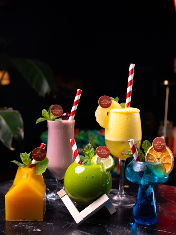 Assorted Colorful Tropical Cocktails And Smoothies Garnished With Fruit And Striped Straws On A Dark Bar.